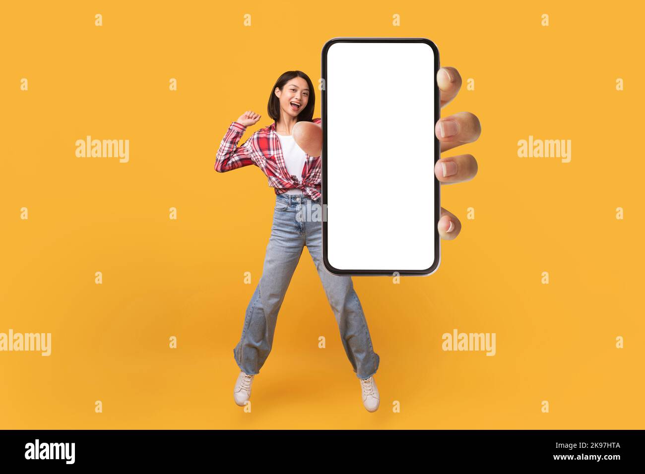 Bonne dame coréenne montrant l'écran vide du smartphone tout en sautant sur fond jaune studio, collage, maquette Banque D'Images