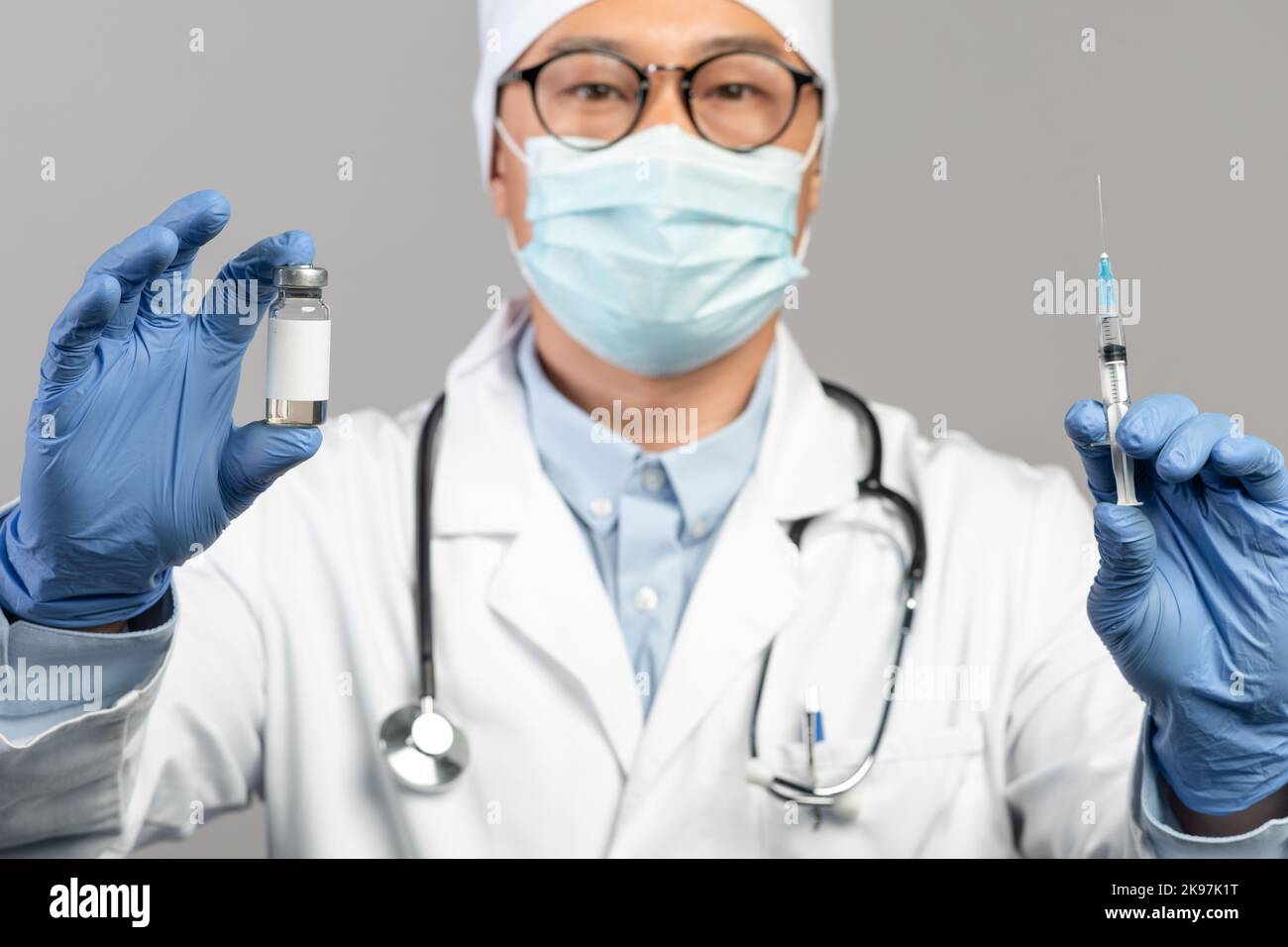 Calme mature chinois médecin en manteau blanc, gants de protection, masque et verres montrent le vaccin et la seringue Banque D'Images