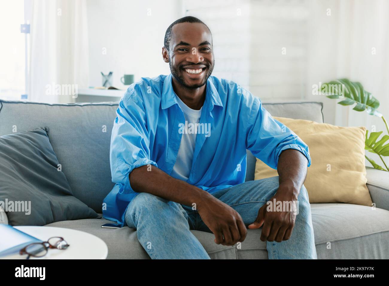 Portrait d'un beau homme noir souriant et regardant l'appareil photo, assis sur un canapé à la maison, passant du temps à l'intérieur Banque D'Images