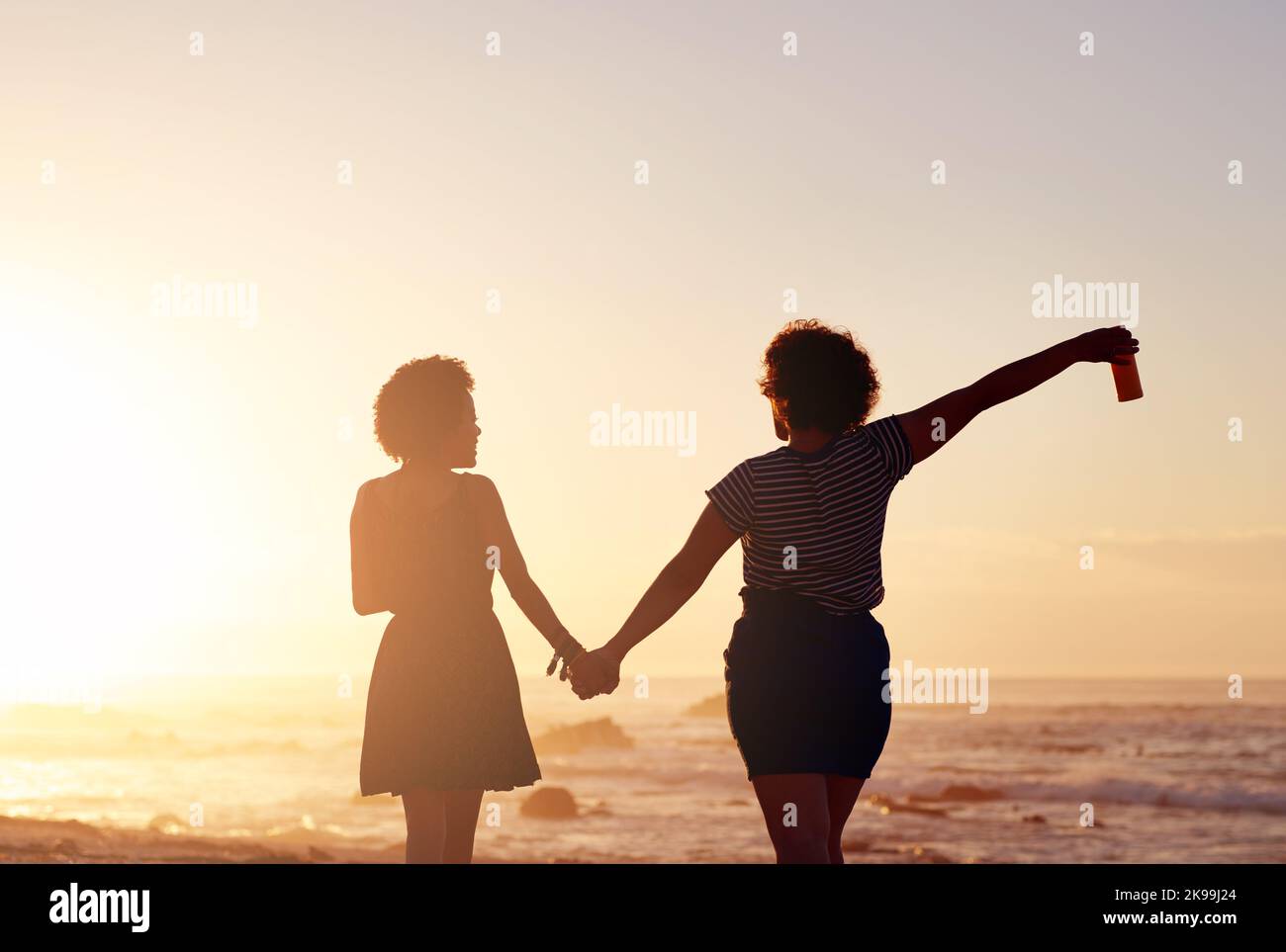 Les amis sont des gens qui aiment la vie ensemble. Vue arrière de deux jeunes femmes méconnaissables se tenant les mains et marchant le long de la plage au coucher du soleil. Banque D'Images