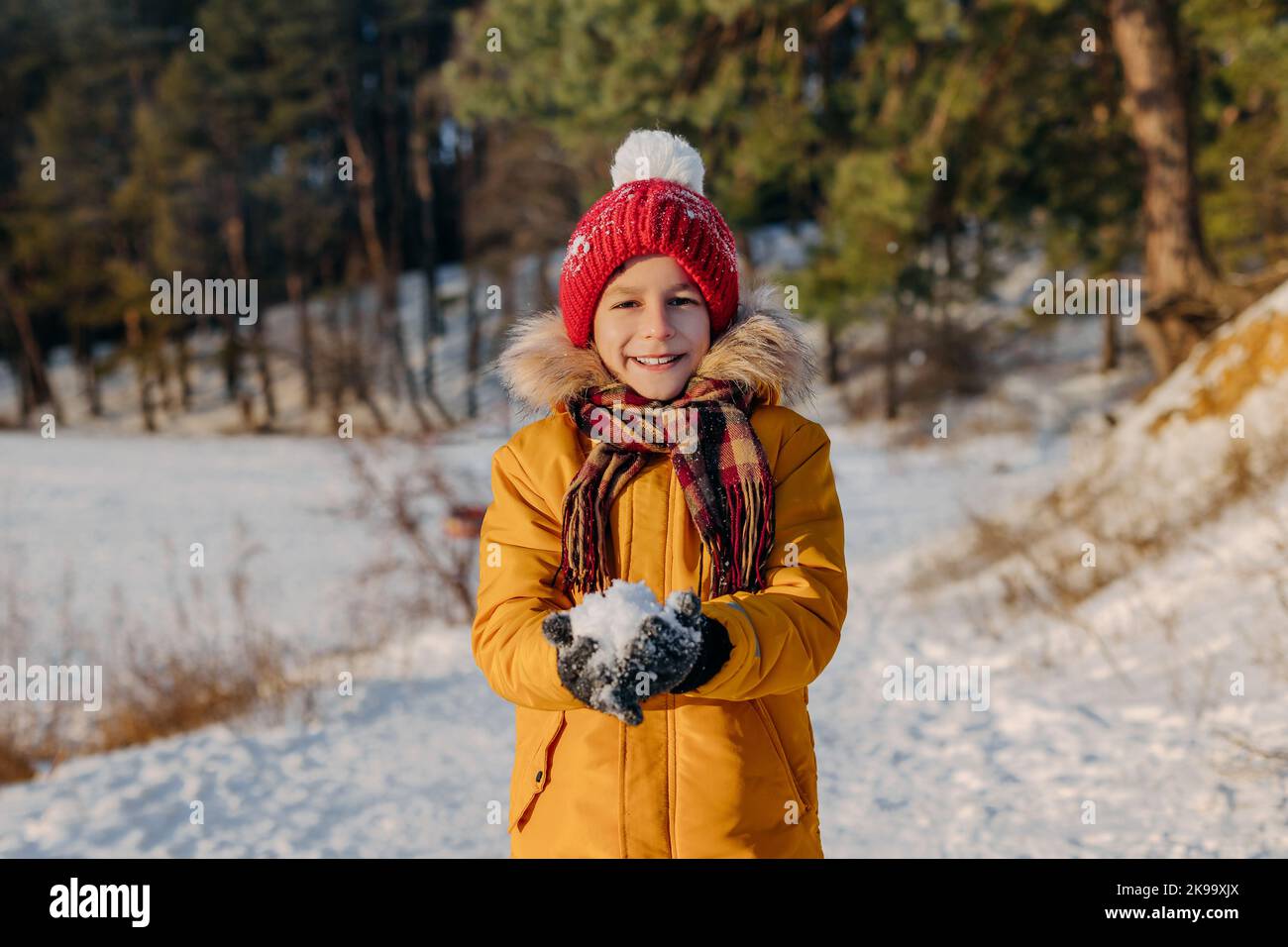 Joyeux enfant tenant dans les mains boule de neige sur le fond de forêt enneigée. Garçon souriant dans des vêtements chauds ayant du plaisir à l'extérieur. Concept des vacances de noël Banque D'Images