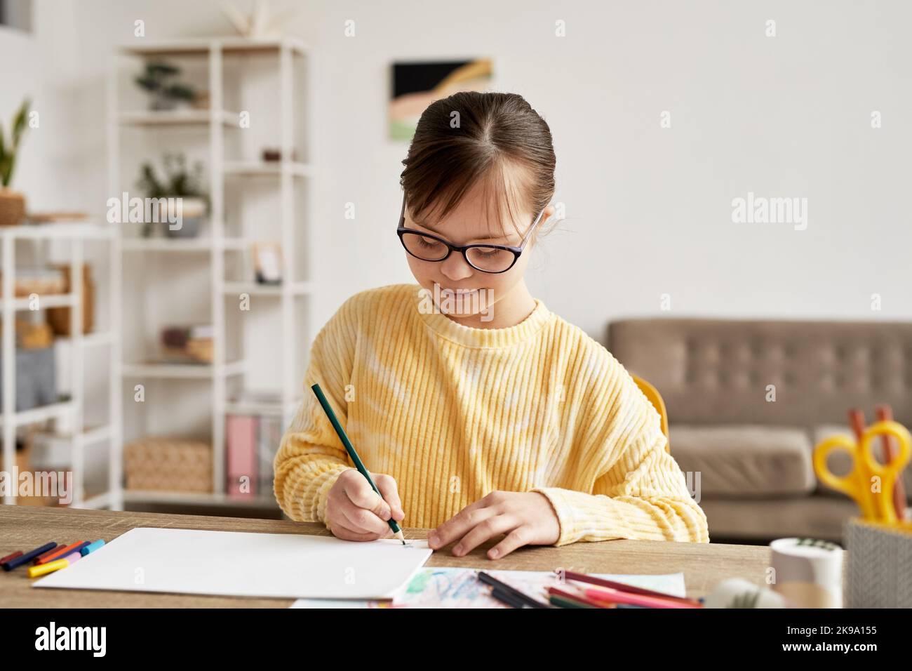 Portrait d'une adolescente atteinte du syndrome de Down, en train de dessiner des images à table dans une pièce confortable et en souriant, espace de copie Banque D'Images