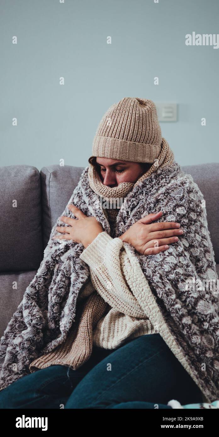 Femme qui gèle à la maison, portant une couverture épaisse, un chapeau d'hiver et une écharpe Banque D'Images