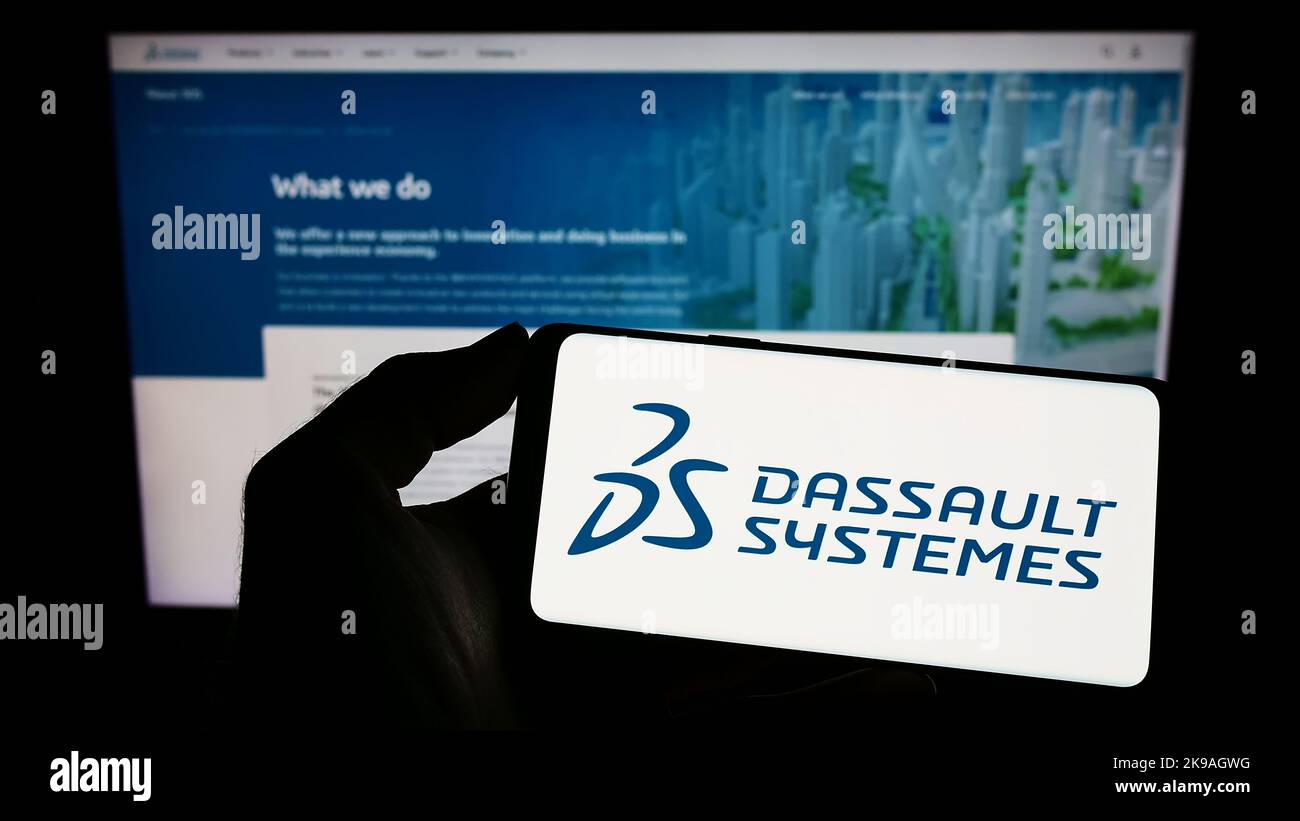 Personne tenant un téléphone portable avec le logo de la société de logiciels française Dassault systèmes se à l'écran en face de la page web d'affaires. Mise au point sur l'affichage du téléphone. Banque D'Images