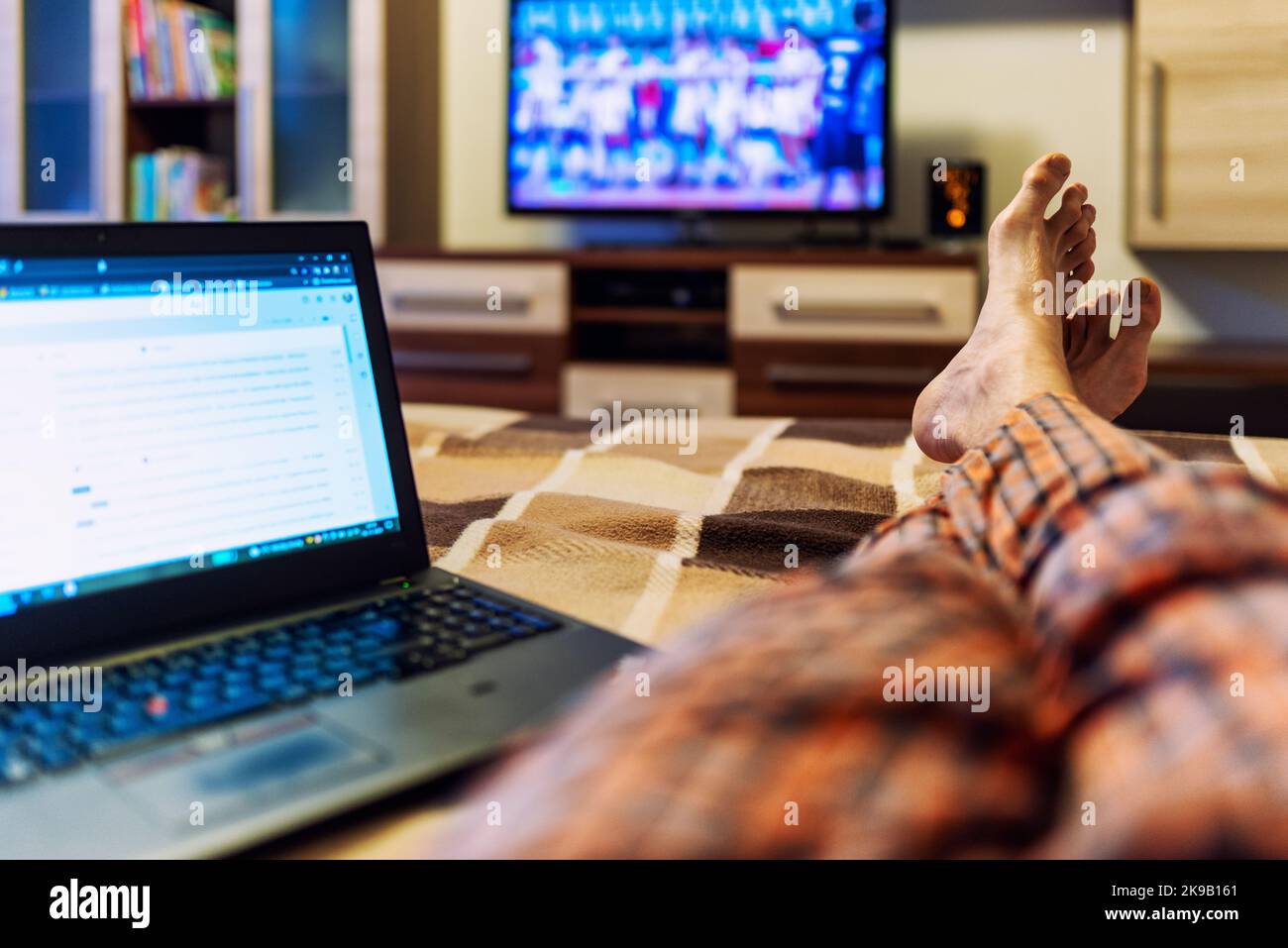 Regarder la télévision et l'écran de l'ordinateur portable dans le salon avec les jambes croisées couchée sur le lit en raison de la quarantaine Banque D'Images