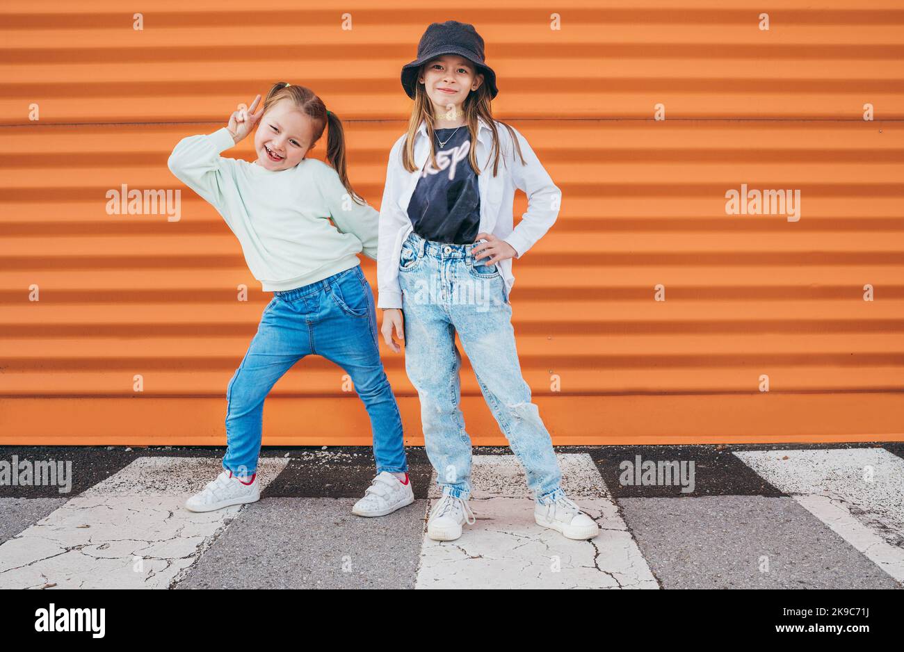 Deux petites filles sœurs vêtues d'enfants mode vêtements de style vogue posant à la caméra sur la route piétonne asphaltée de la ville près de l'arrière-plan du mur orange Banque D'Images