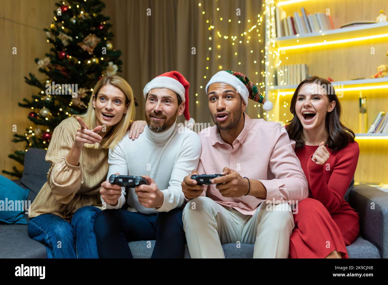 Groupe de quatre amis du nouvel an se détendant et célébrant les fêtes de Noël, les invités assis sur un canapé, hommes et femmes, jouant à des jeux vidéo sur des consoles de joystick. Banque D'Images