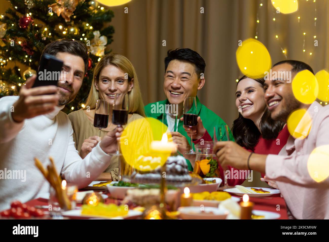 Une compagnie de personnes interraciales célébrant ensemble le nouvel an, assis à la table des fêtes. L'homme tient le téléphone, tout le monde parle lors d'un appel vidéo avec des amis, salutation, sourire. Banque D'Images