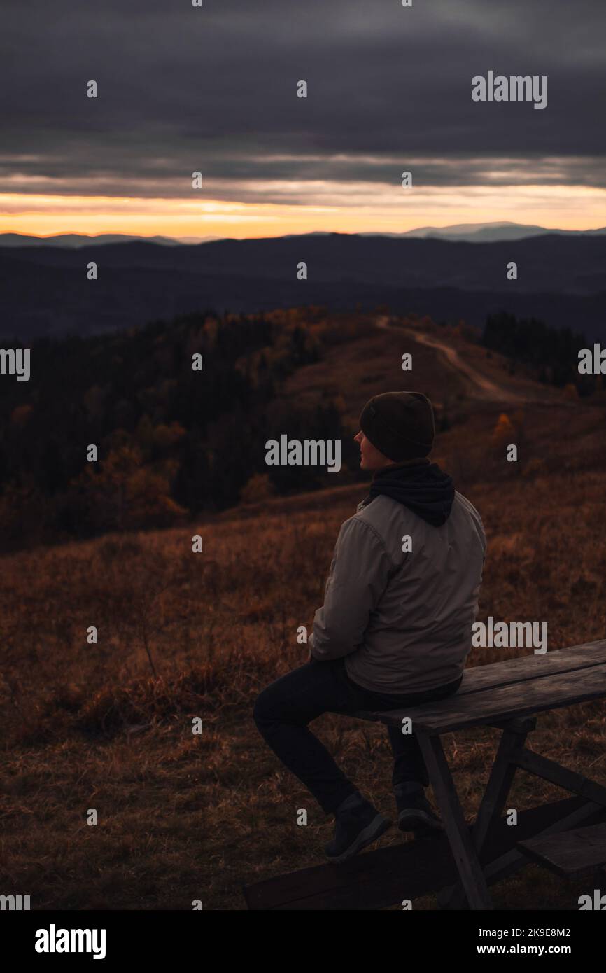 Le jeune homme est assis sur le banc et regarde la vue sur les montagnes au coucher du soleil Banque D'Images