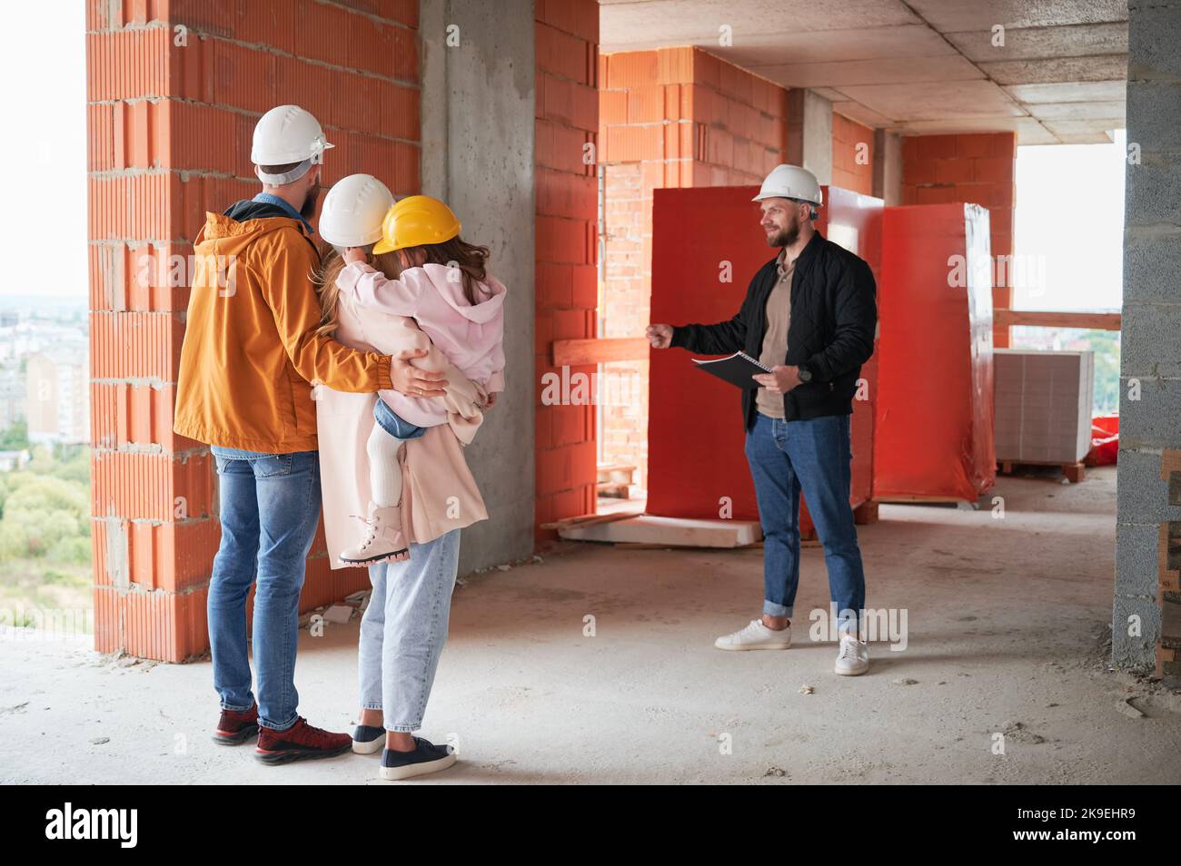 Homme et femme avec enfant debout à l'intérieur de l'immeuble d'appartements en construction et parlant avec le constructeur. Famille futurs propriétaires discutant du processus de construction avec le travailleur de la construction. Banque D'Images