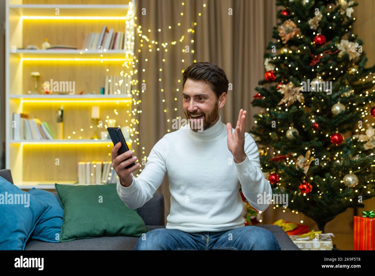 Un homme parle lors d'un appel vidéo à la maison avec un téléphone, s'assoit sur un canapé près d'un arbre de Noël, enregistre les messages d'accueil vidéo du nouvel an et des vacances de Noël. Banque D'Images