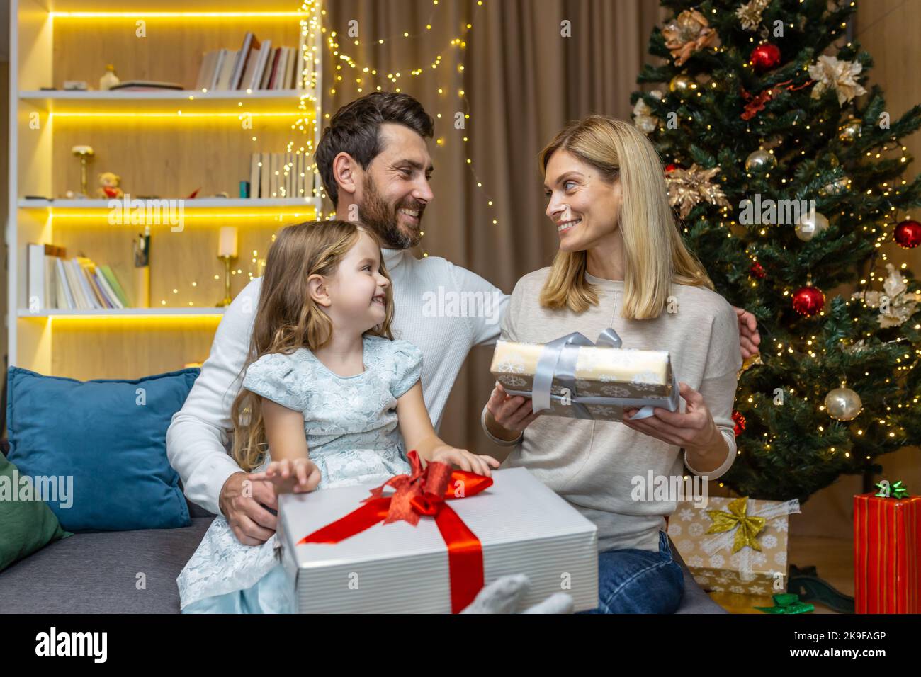 Nouvel an et vacances de Noël, famille heureuse avec fille échangeant des cadeaux et saluant les uns les autres assis sur le canapé près de l'arbre de Noël à la maison. Banque D'Images