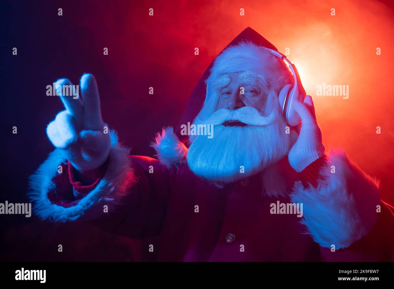 Le Père Noël montre 2 doigts qui écoutent de la musique sur un casque lors d'une fête. Lumière néon bleu rouge et fumée. Banque D'Images