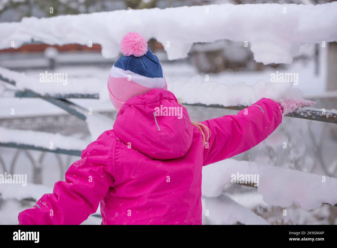 Une petite fille dans un costume de ski rose vif fait tomber la neige de la main courante d'un pont. Le plaisir des enfants. Banque D'Images