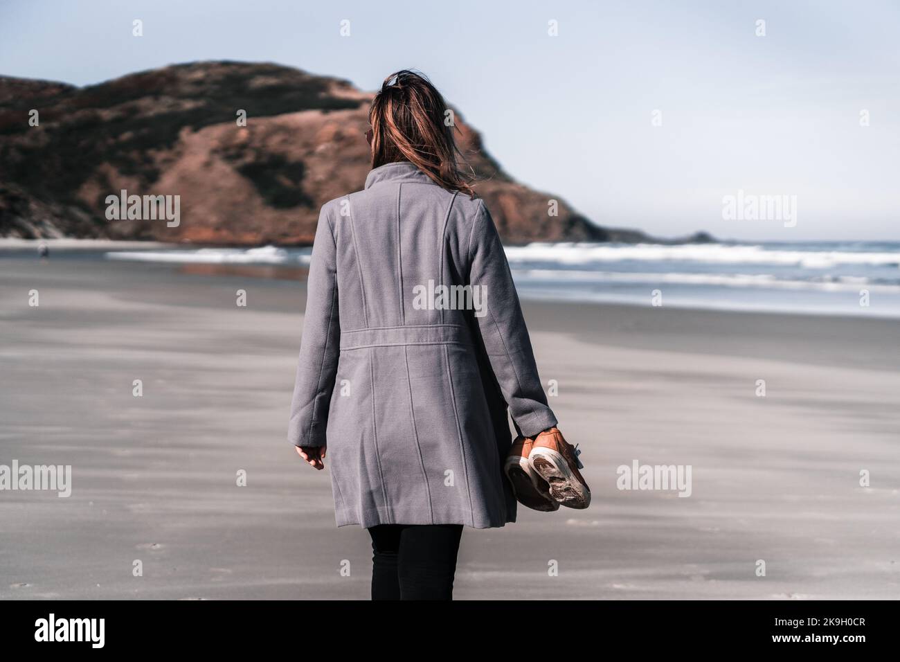 jeune femme caucasienne marchant sur la plage sable calme et détendu portant un long manteau gris pantalon noir et des baskets brunes dans sa main, sandfly bay, nouveau Banque D'Images