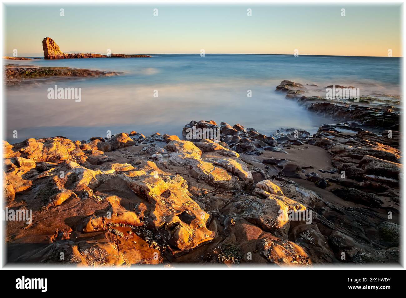 Une scène côtière sereine au coucher du soleil avec des rochers accidentés recouverts de mousse, baignés d'une lumière chaude et dorée contrastant avec une eau douce rêveuse en arrière-plan. Banque D'Images