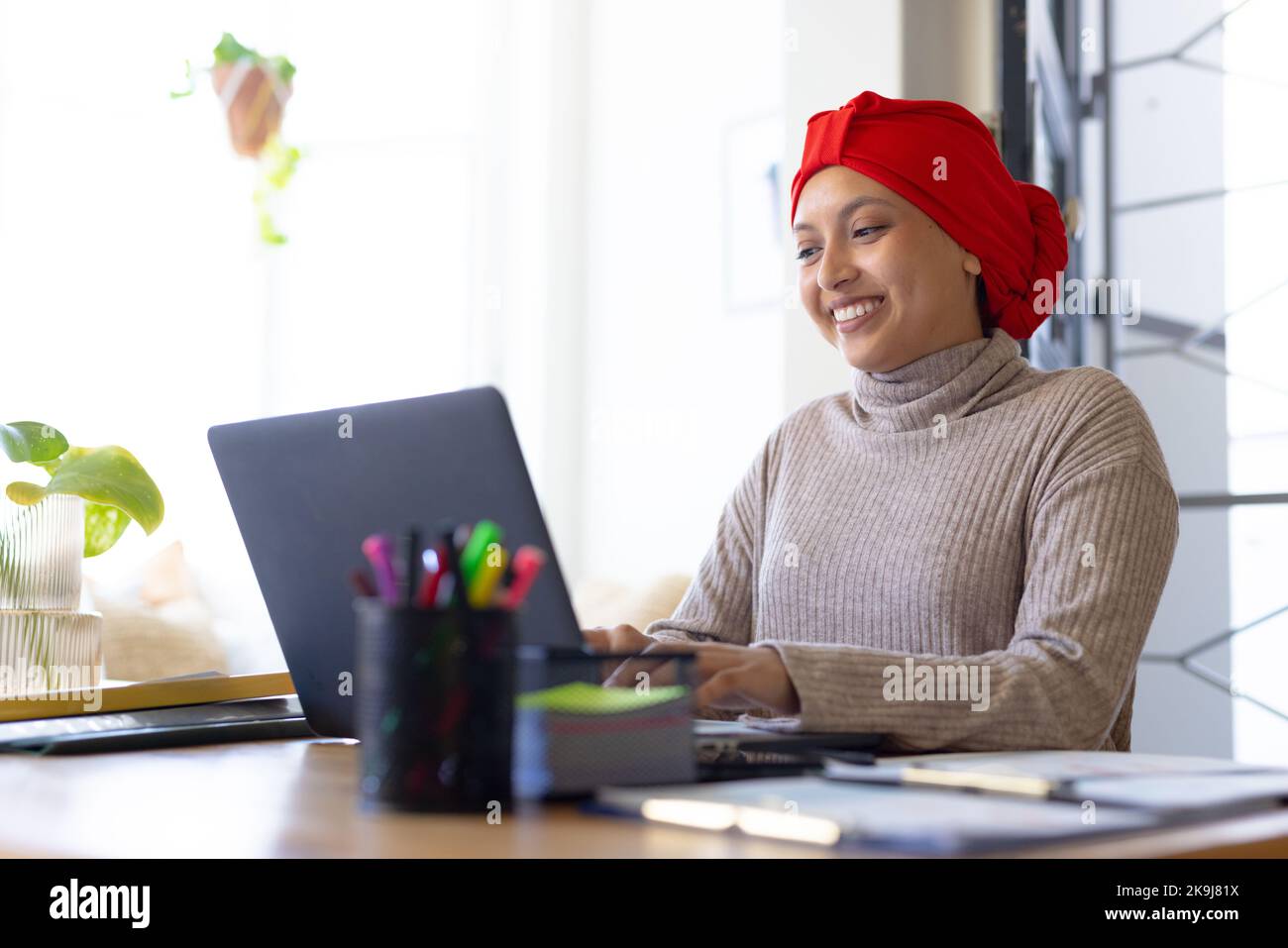 Heureuse femme biraciale portant le hijab, assise à une table et utilisant un ordinateur portable Banque D'Images