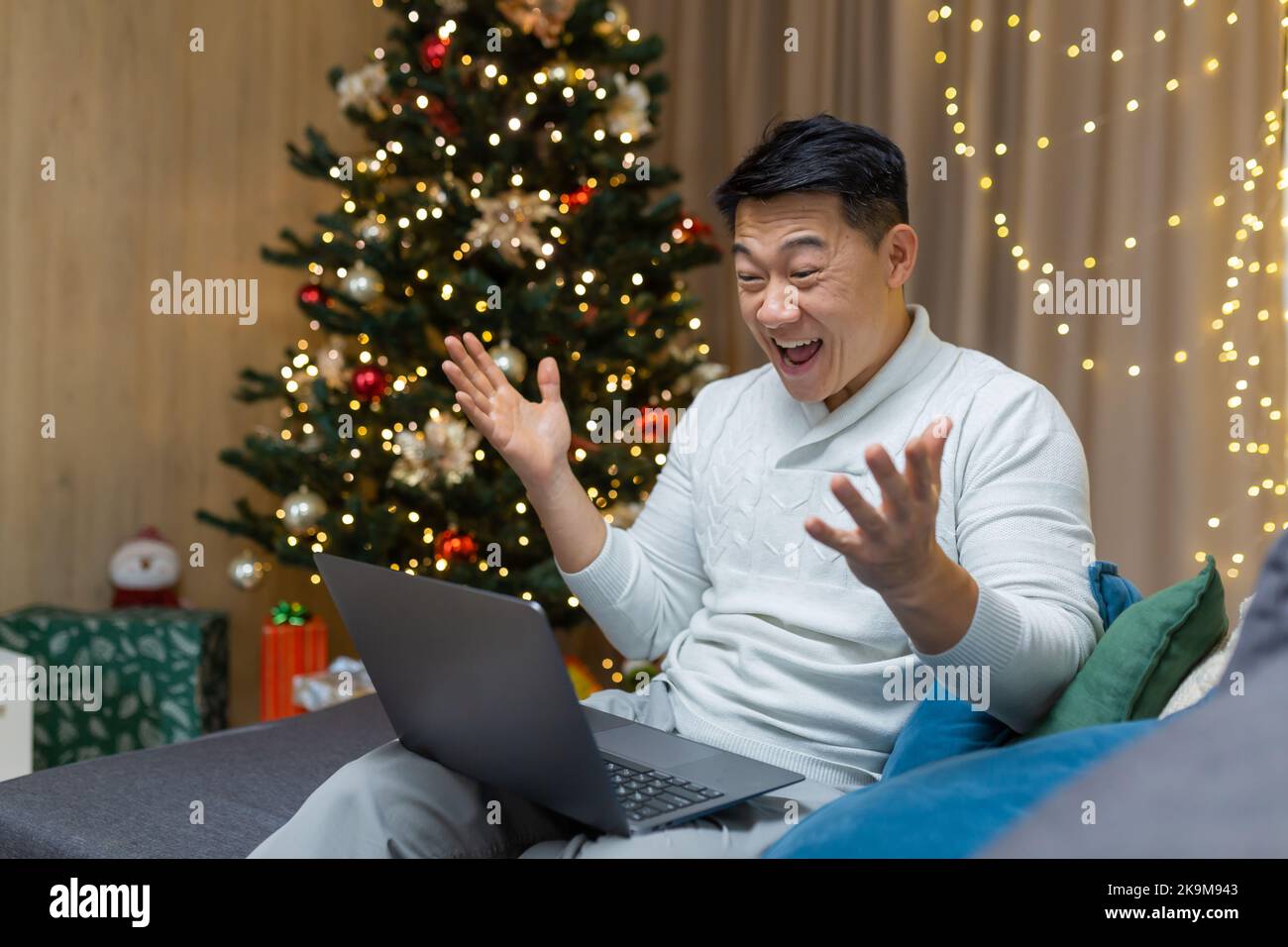 Homme asiatique lisant les bonnes nouvelles sur ordinateur portable à la maison pour Noël, homme d'affaires travaillant à distance pendant les vacances du nouvel an assis sur un canapé dans le salon, célébrant le succès de la victoire. Banque D'Images