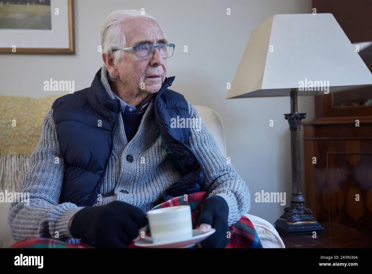 Homme senior portant des vêtements supplémentaires avec boisson chaude essayant de rester au chaud à la maison en crise d'énergie Banque D'Images