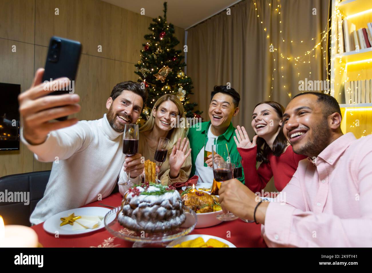 Divers amis célébrant Noël et le nouvel an dans la soirée, assis à la table, boire du vin et manger, prendre un selfie ensemble au téléphone près de l'arbre de Noël. Banque D'Images