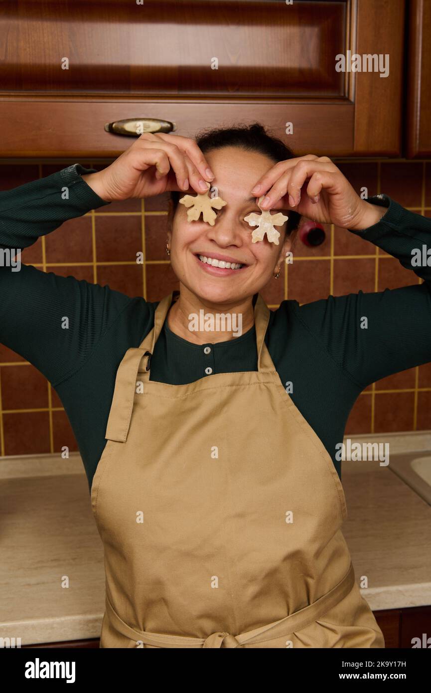 Femme de ménage gaie tenant deux pâtes de pain d'épice en forme de flocons de neige près de ses yeux et souriant un magnifique sourire Banque D'Images