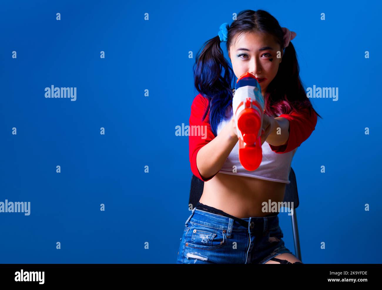 Jeune femme dans Harlequin Costume assis et visant un pistolet à eau à la caméra avec un espace de copie à gauche dans un arrière-plan bleu isolé Banque D'Images