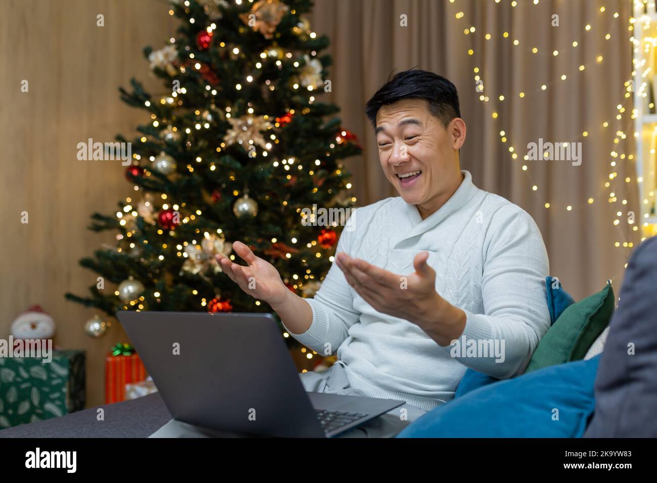 Joyeux noël homme faisant des appels vidéo à l'aide d'un ordinateur portable assis sur un canapé dans le salon à la maison, homme asiatique saluant amis en ligne joyeux nouvel an souriant et parlant heureusement. Banque D'Images
