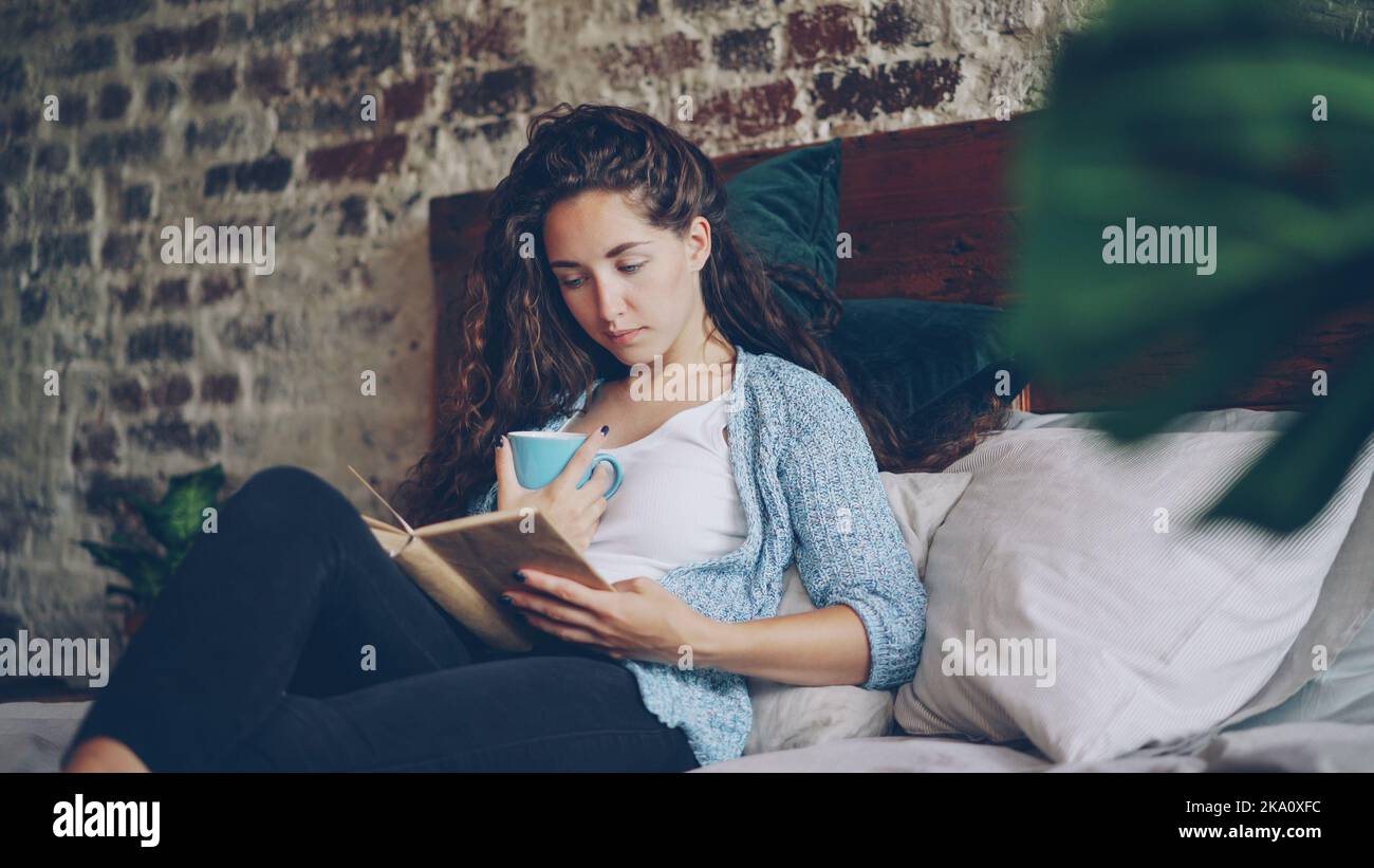 Une fille gaie est en train de lire un livre drôle dans la chambre, allongé  sur un lit double confortable, souriant et tenant une tasse avec un verre.  Passe-temps, gens heureux, beaux