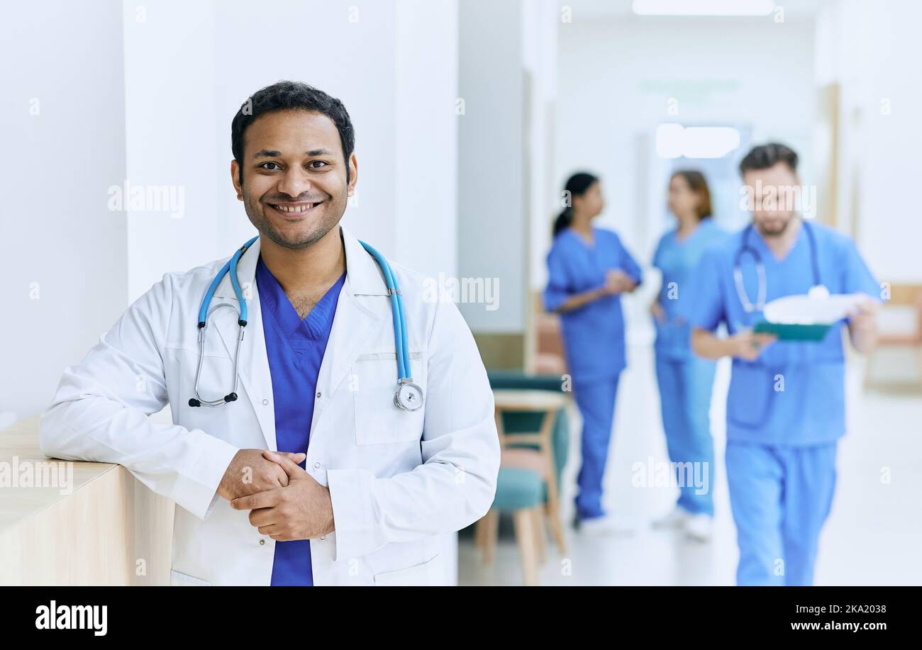 Médecin indien souriant debout dans le couloir de l'hôpital moderne sur fond d'assistants médicaux et d'infirmières travaillant. institution médicale Banque D'Images