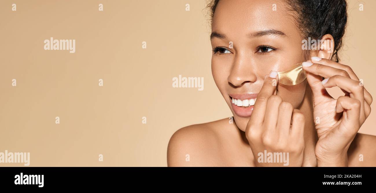 Souriante femme afro-américaine applique des taches hydratantes dorées sous ses yeux pour les cercles sombres et la puffiness, sur fond pastel Banque D'Images