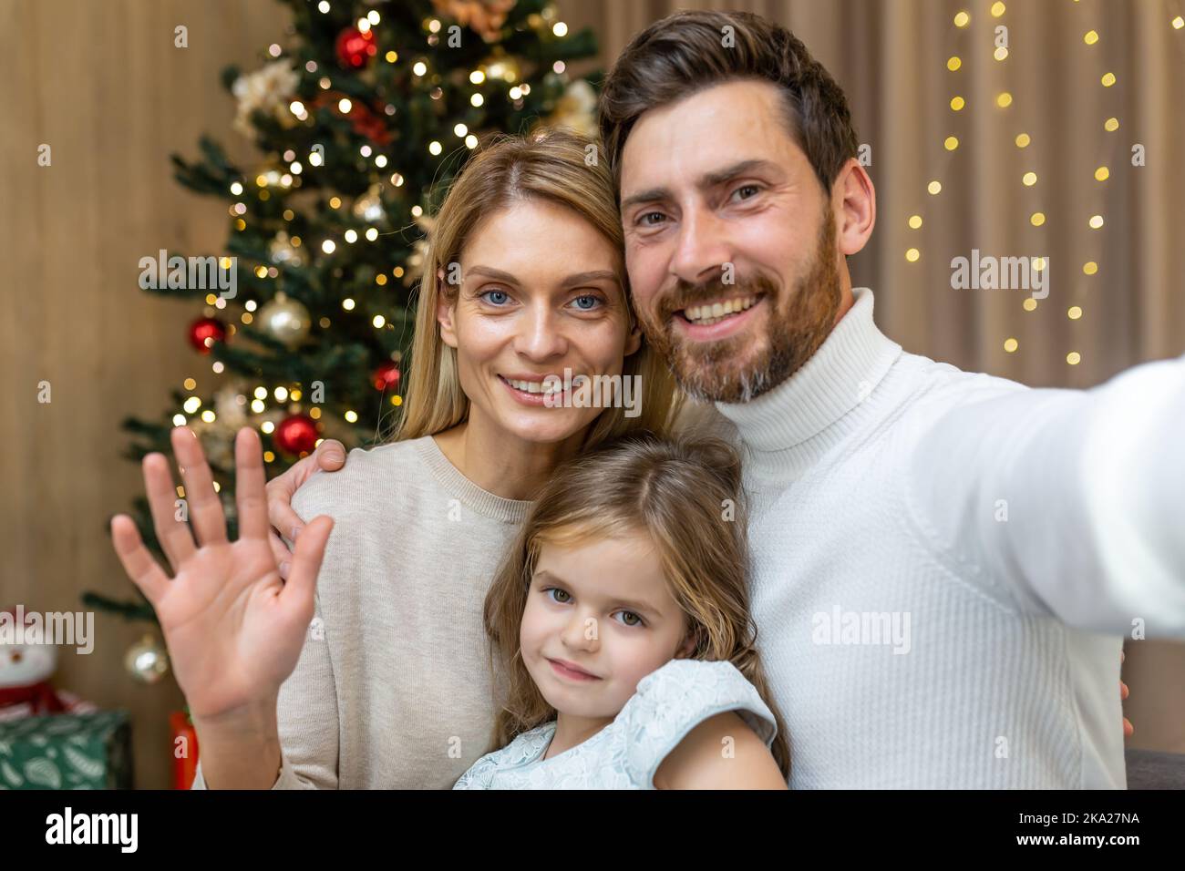 Portrait de fête d'une jeune famille heureuse près de l'arbre de Noël à la maison. Maman, papa et fille prennent un selfie, regardez l'appareil photo, sourire, vague. Banque D'Images