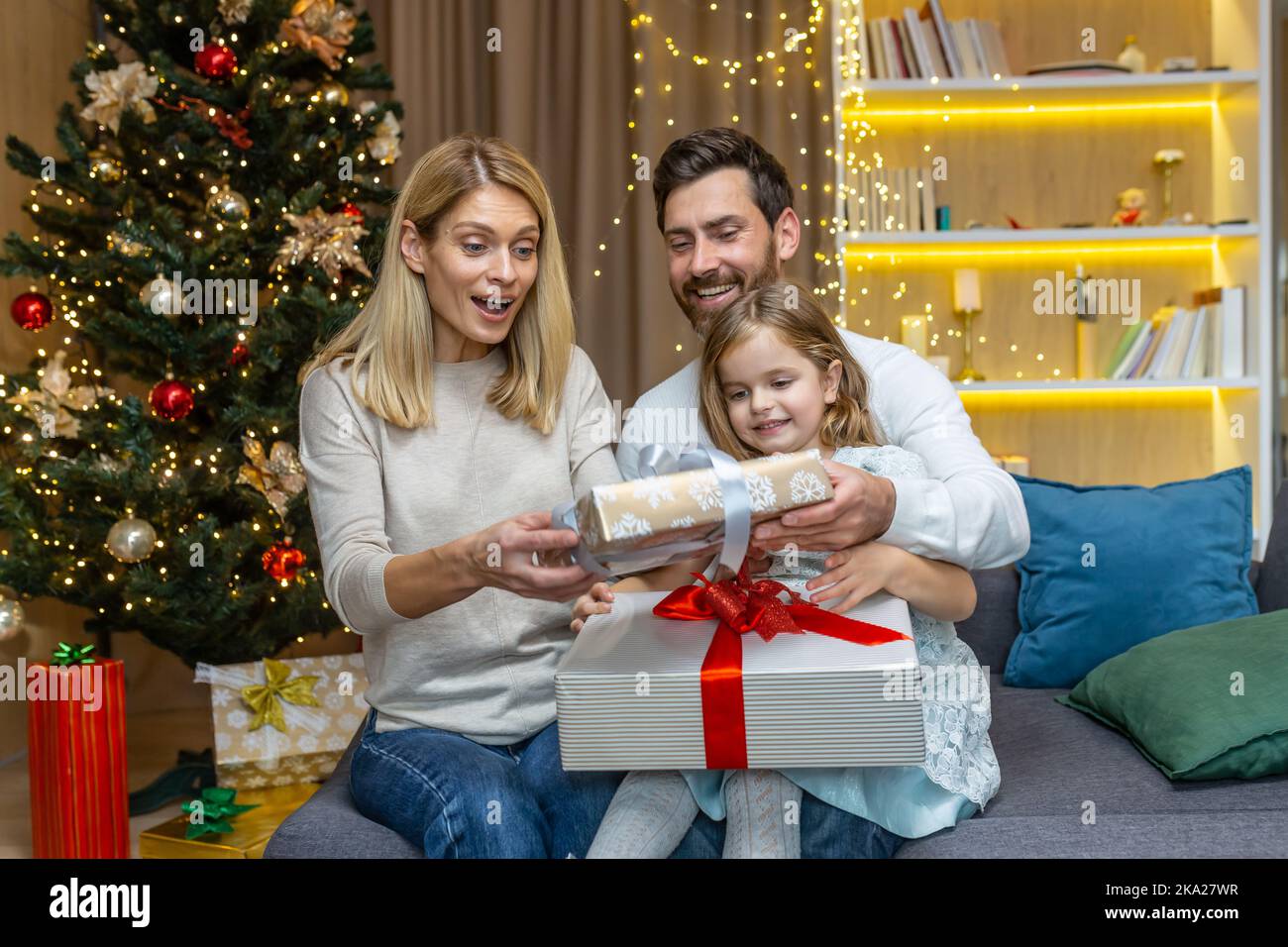 Portrait de fête d'une jeune famille heureuse près de l'arbre de Noël à la maison. Maman, papa et petite fille sont assis sur le canapé et échangent des cadeaux, en se donnant joyeusement les autres boîtes avec des surprises. Banque D'Images