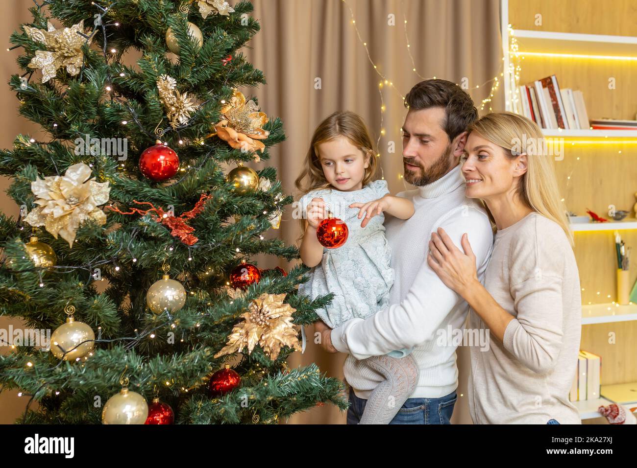 Une jeune famille heureuse décorera un arbre de Noël pour les vacances de Noël. Papa tient sa fille dans ses bras, la fille porte un jouet, la maman embrasse le papa. Banque D'Images