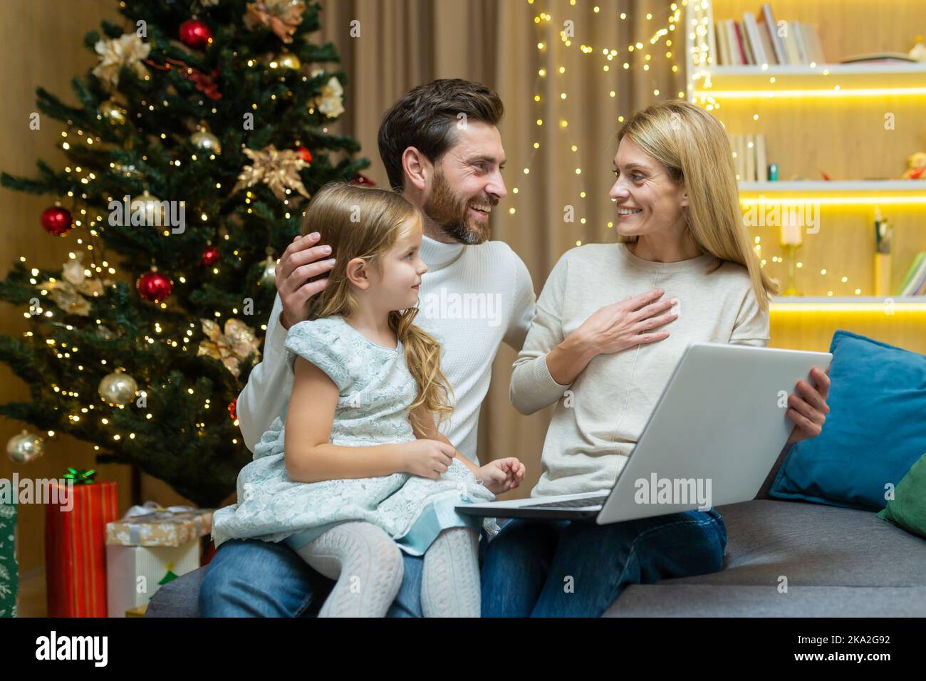 Les parents matures enseignent à leur enfant d'utiliser un ordinateur portable étudiant l'apprentissage en ligne de la navigation sur Internet ensemble, assis sur le canapé dans le salon arrière-plan arbre de Noël père et mère avec enfant fille. Banque D'Images