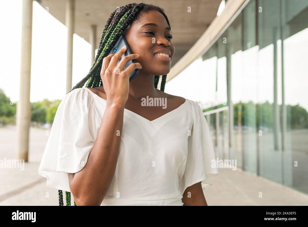 Content jeune femme afro-américaine avec des cheveux tressés verts parlant au-dessus de smartphone tout en étant debout dans la rue Banque D'Images