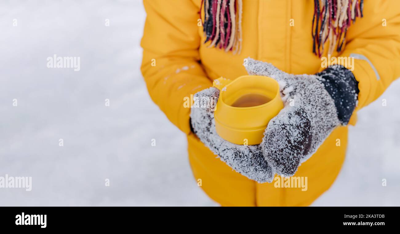 Gros plan des mains tenant une tasse de thé chaud jaune sur fond de neige. Garçon en gants de neige tricotés et écharpe avec mug chaud. Thème des vacances d'hiver avec Banque D'Images