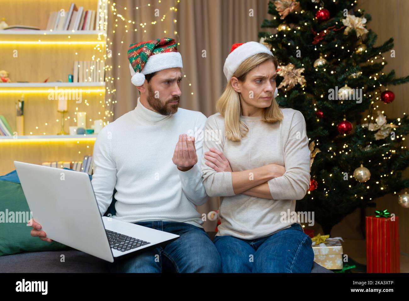 Un homme piquant ne permet pas à une femme à la maison de faire des achats dans le magasin en ligne de shopping, un couple marié, un homme et une femme, sont assis sur le canapé à la maison près de l'arbre de Noël dans les chapeaux du nouvel an, les vacances du nouvel an. Banque D'Images