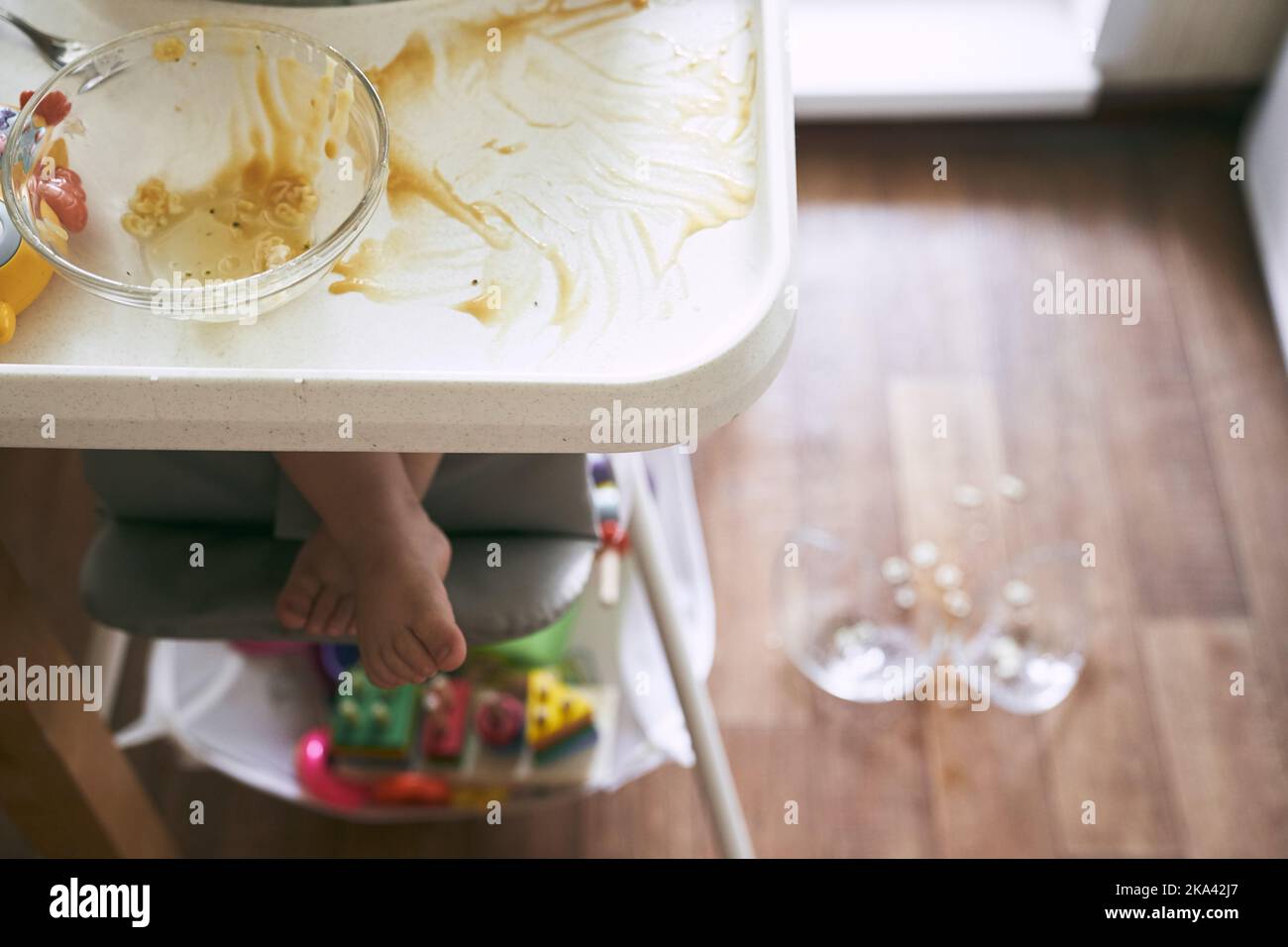 Un petit bébé sur une chaise haute a déposé une tasse de soupe de nouilles sur le parquet. La plaque transparente est divisée en deux parties. Nouilles sous forme de chiffres. La table est sale. Photo de haute qualité Banque D'Images