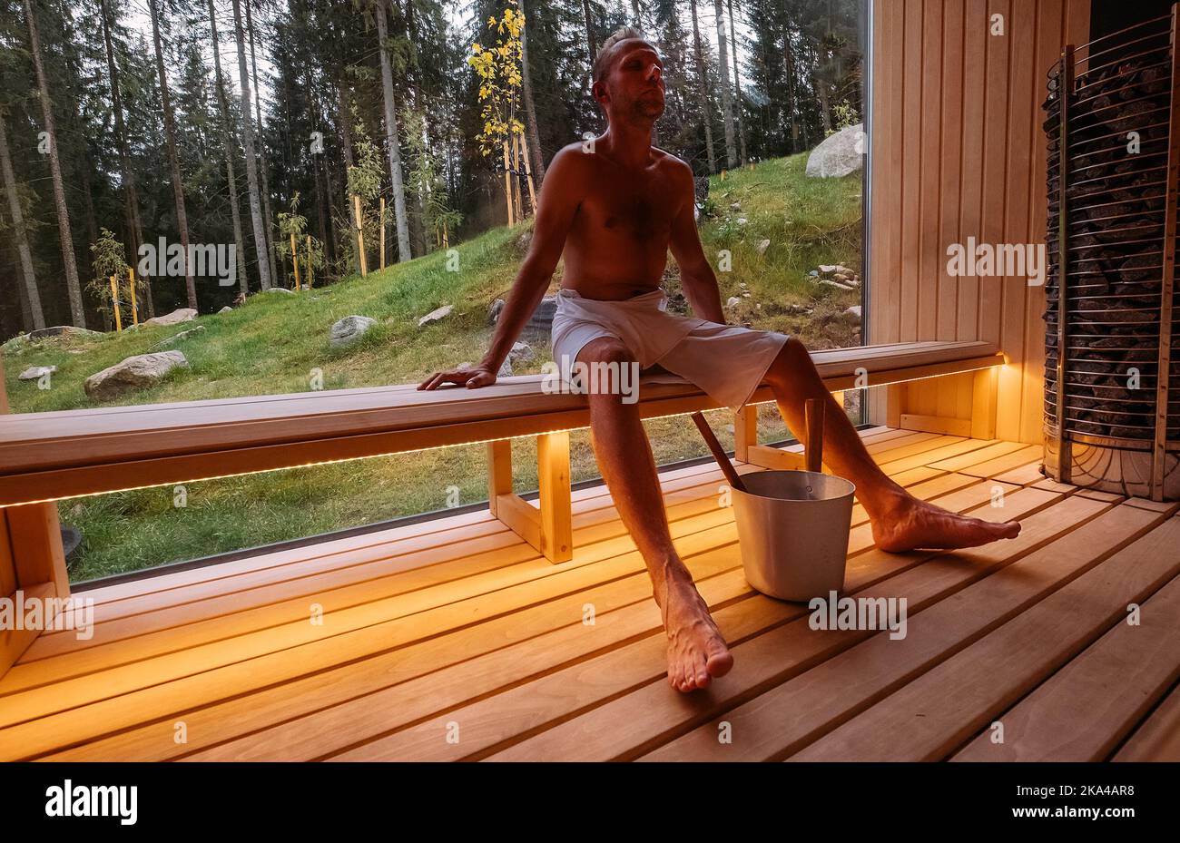 Serviette blanche enveloppée d'un homme reposant sur un banc en bois dans un sauna finlandais chaud avec une grande fenêtre avec vue sur la forêt verte et agréable Banque D'Images