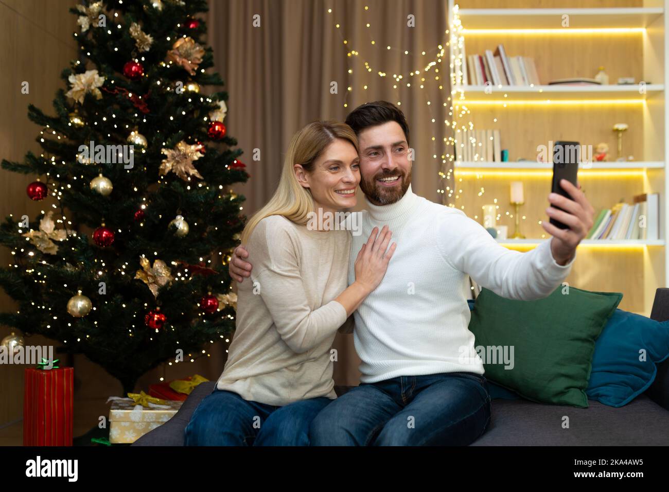 Couple marié homme et femme ensemble sur le canapé à la maison arbre de Noël avec téléphone intelligent parler appel vidéo et prendre selfie, la famille mature saluant des amis en ligne à distance pour le nouvel an et Noël. Banque D'Images