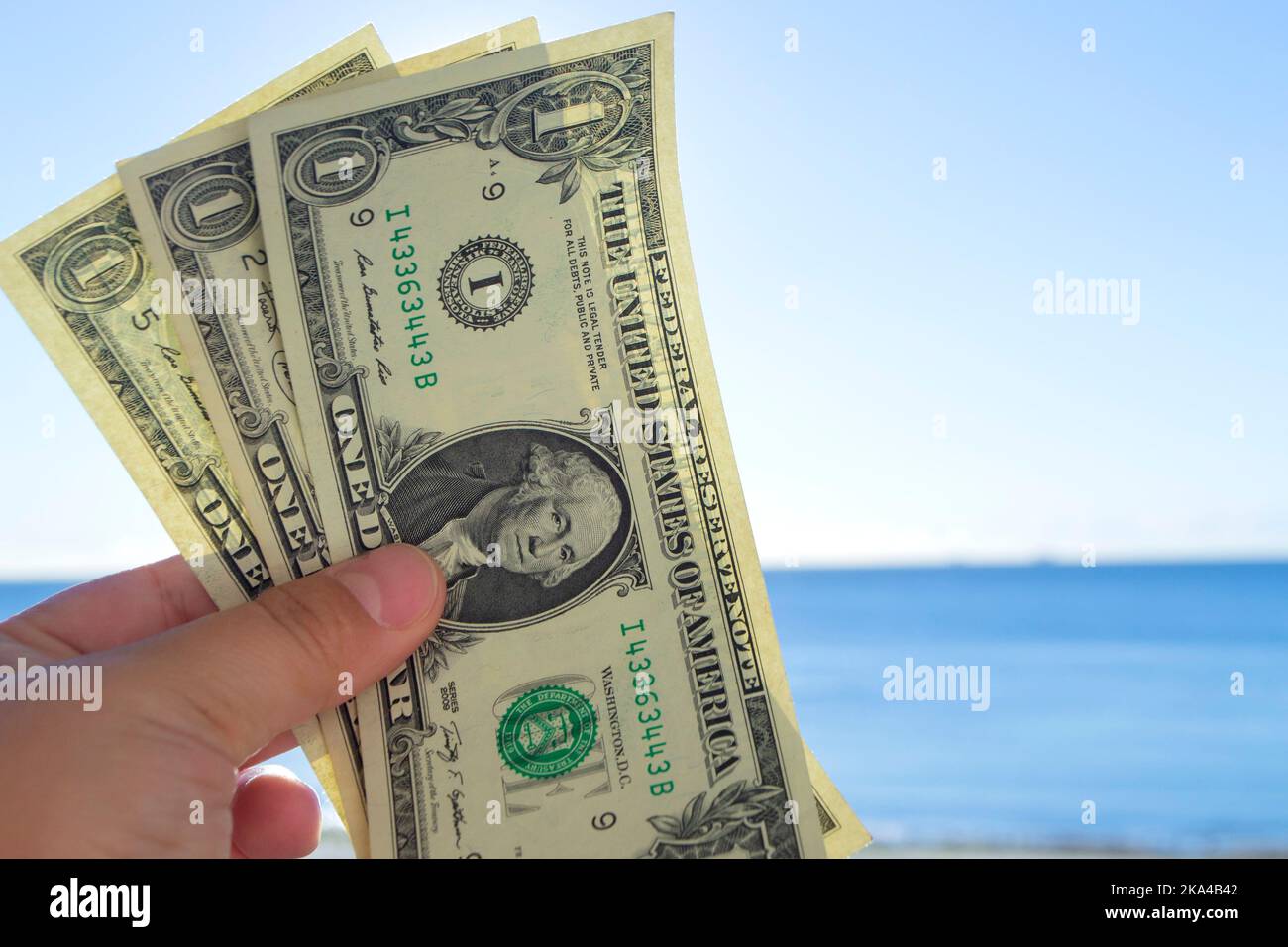 Homme tenant trois billets d'un dollar dans sa main sur fond de mer bleue et de ciel bleu le jour ensoleillé de l'été. Le concept de l'argent, de la finance, de la monnaie, des voyages, du tourisme, vacances, repos Banque D'Images