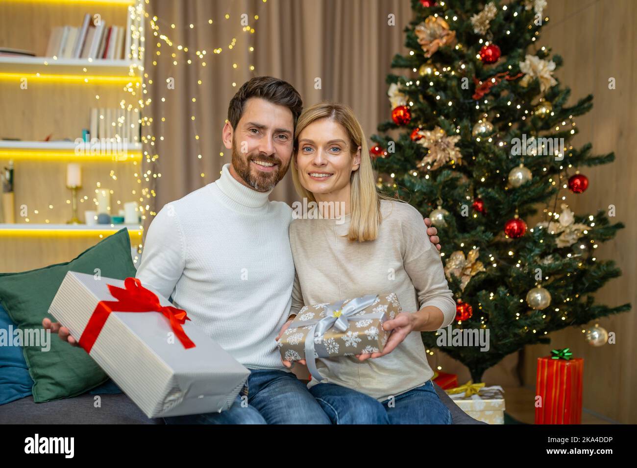 Portrait d'un homme barbu et d'une femme blonde de famille mûre regardant un appareil photo près de l'arbre de Noël, couple célébrant noël et nouvel an avec des cadeaux à la maison assis sur un canapé dans le salon. Banque D'Images