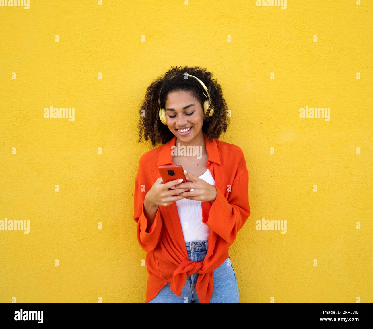 Jeune femme afro-américaine souriante discutant avec un téléphone portable et un casque sur un mur jaune Banque D'Images