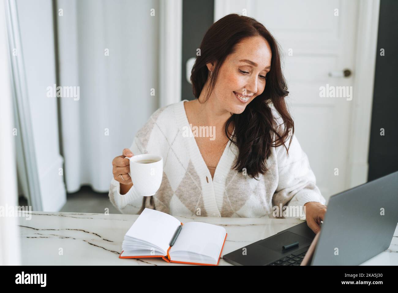 Femme souriante brune avec cheveux longs dans un gilet blanc travaillant sur un ordinateur portable dans un bureau moderne lumineux Banque D'Images
