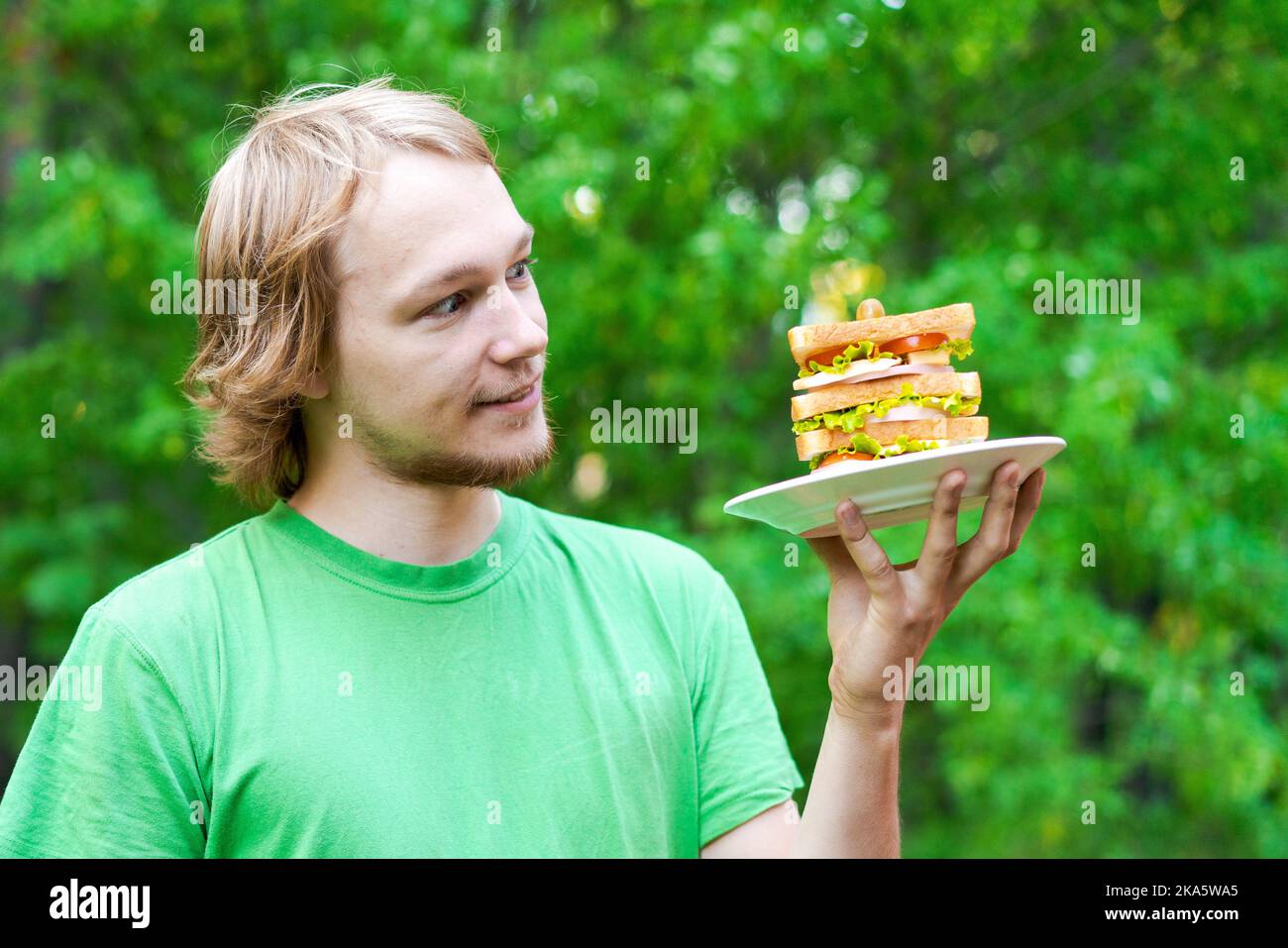 Jeune homme tenant un gros sandwich à la saucisse sur l'assiette. L'étudiant mange de la nourriture rapide dans un paquet sur fond de feuillage vert. Aliments indésirables rapides. Un homme très affamé. Concept de régime. Banque D'Images