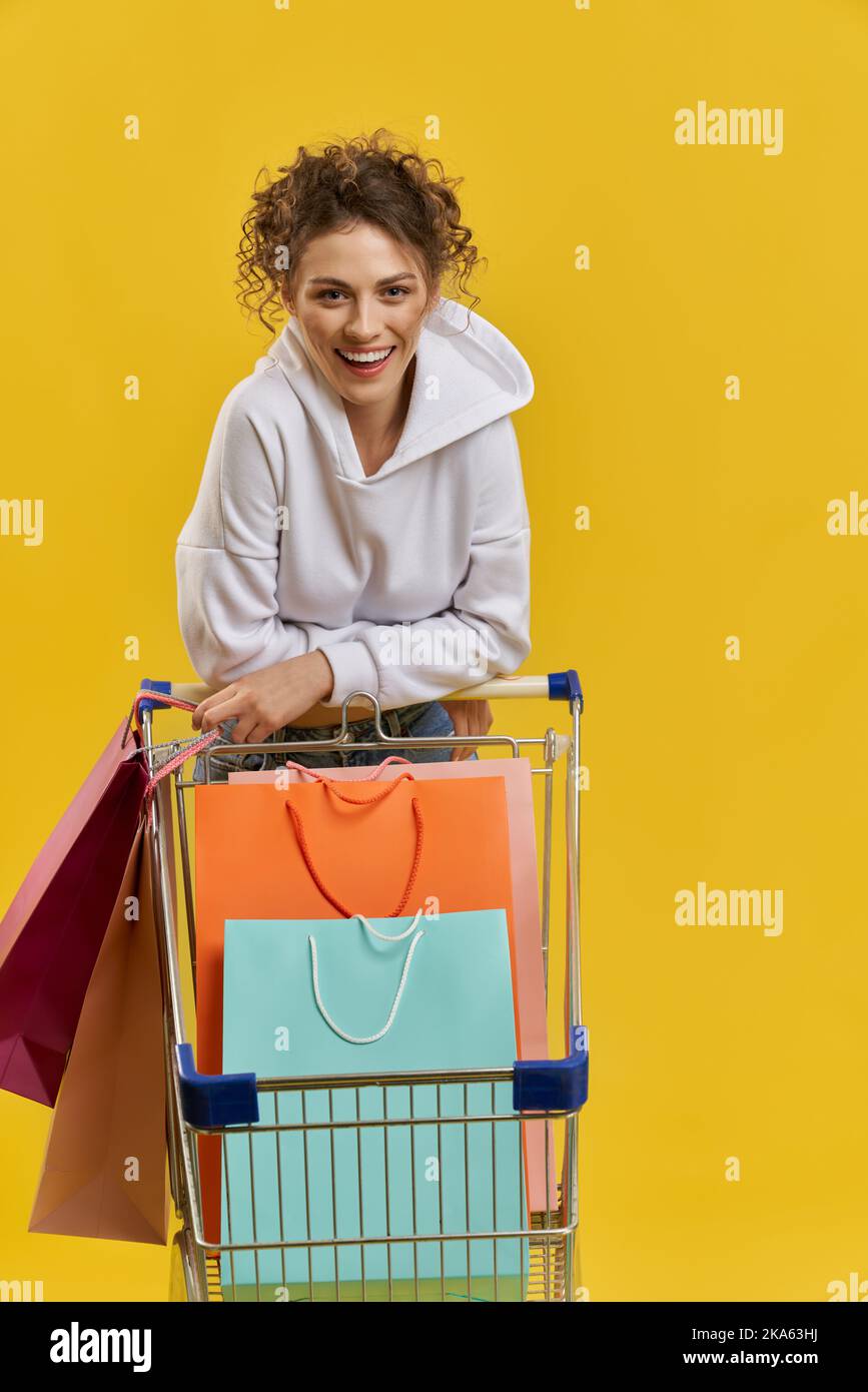 Vue avant de la jolie fille debout avec un chariot, penchée vers l'avant. Belle femme blonde avec des cheveux bouclés faisant du shopping, souriant, regardant l'appareil photo. Isolé sur fond jaune, Banque D'Images Vue avant de la jolie fille debout avec un chariot, penchée vers l'avant. Belle femme blonde avec des cheveux bouclés faisant du shopping, souriant, regardant l'appareil photo. Isolé sur fond jaune, Banque D'Images