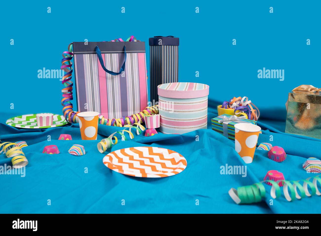 Table de fête désordonnée avec sacs et boîtes cadeaux, banderoles, assiettes et tasses en plastique sur une table avec une nappe bleue sur un fond bleu clair Banque D'Images