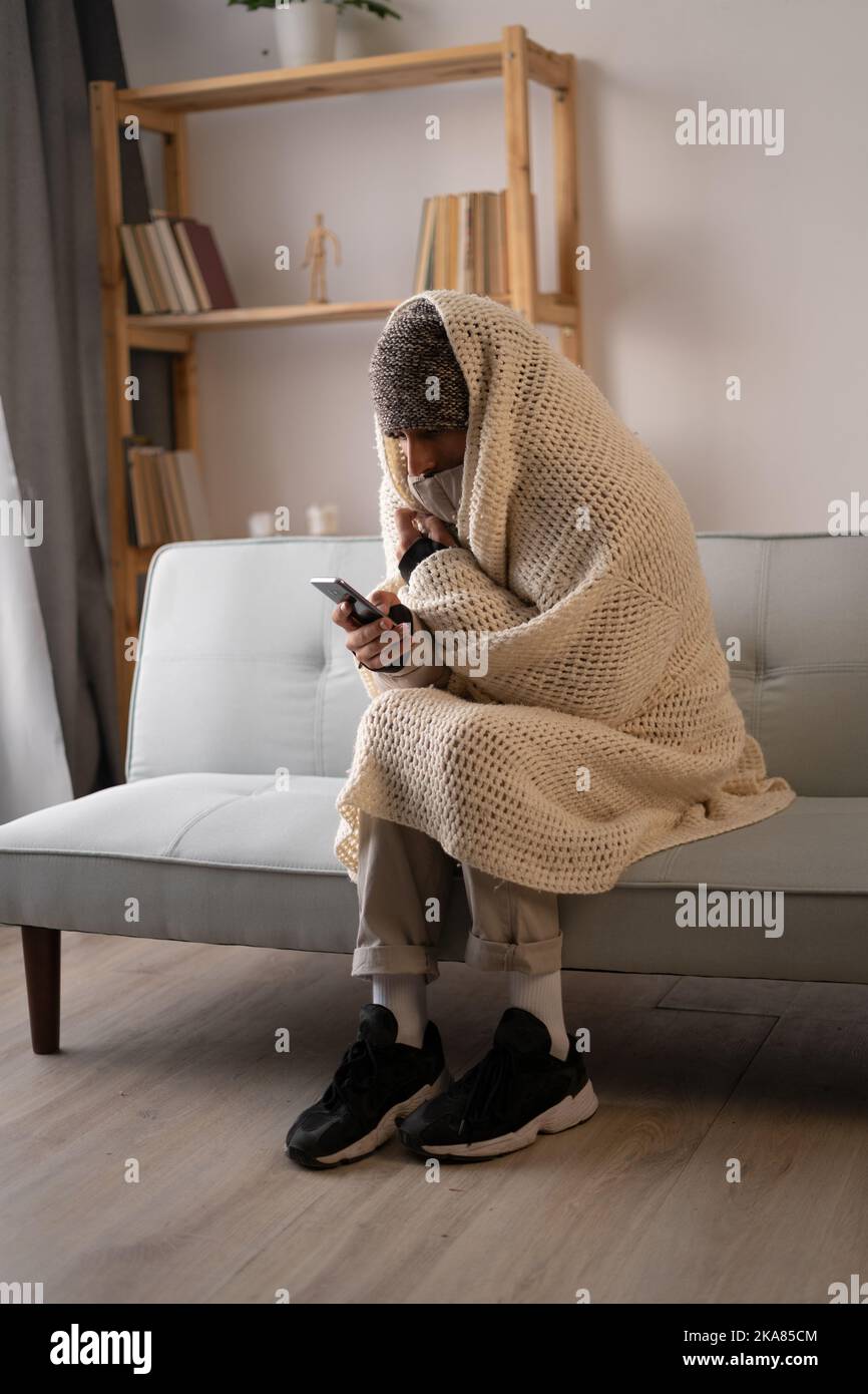 Jeune homme habillé de vêtements d'hiver se sentant froid assis à la maison sans chauffage, jouant sur smartphone. Pas de chauffage Banque D'Images