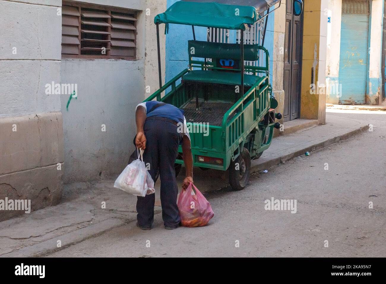 Un Afro-Caribbean ramasse un sac en plastique rempli de produits après avoir pris une pause. Un tricycle électrique Minerva est stationné sur le trottoir. Banque D'Images