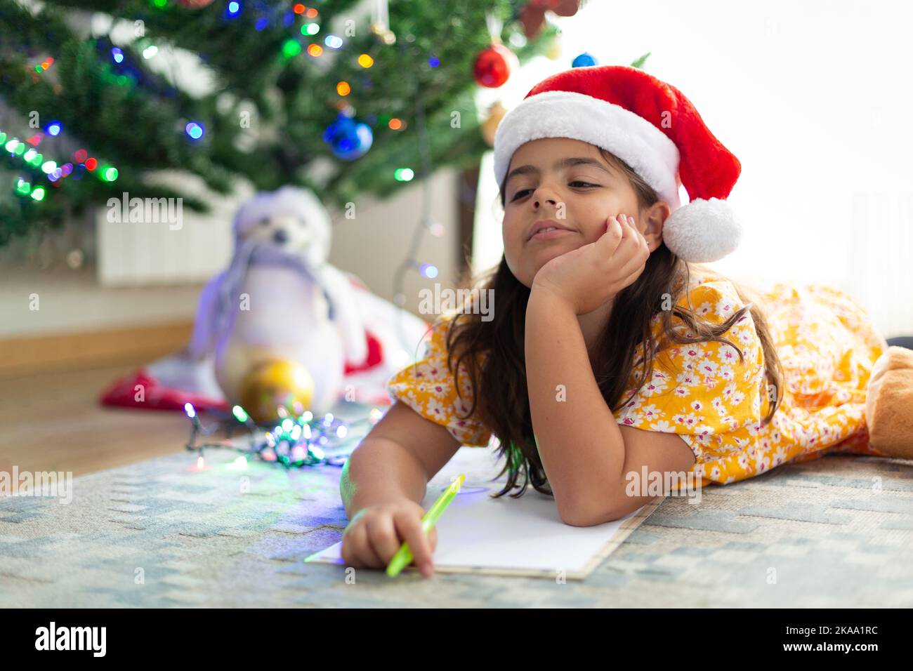Bonne petite fille caucasienne pensant à la liste de souhaits de Noël. Elle est à la maison allongée sur le tapis. Banque D'Images
